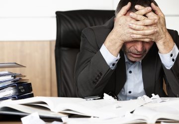 Worried businessman sitting at office desk full with books and papers being overloaded with work.
